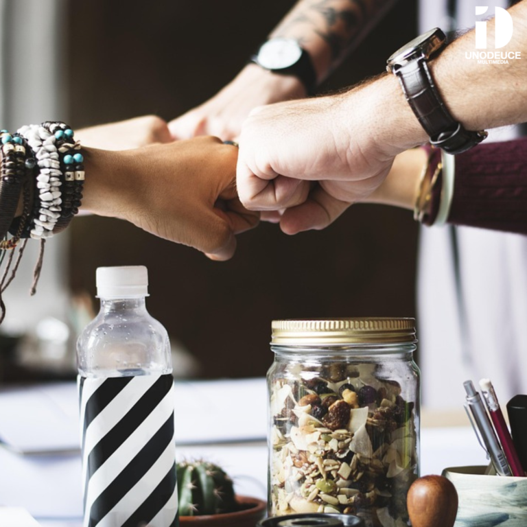 Four hands—representing diverse individuals—meet in a fist bump above a table. On the table are a striped water bottle, a glass jar of mixed nuts and dried fruit, a small potted cactus, and pens in a holder. In the top right corner, the “UnoDeuce Multimedia” logo appears.