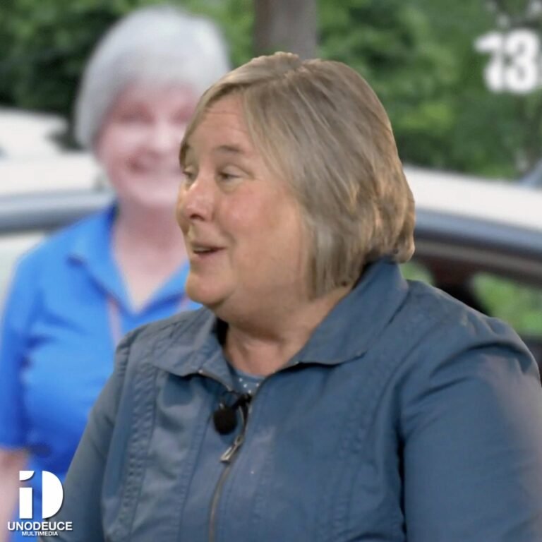 **Alt Text:** A woman with short blonde-gray hair, wearing a blue jacket and lapel microphone, smiles while speaking during an on-camera interview. In the background, a blurred image of the same woman in a blue shirt stands near a car. The UnoDeuce Multimedia logo appears in the bottom left corner.