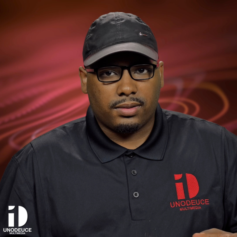 A man sporting a black baseball cap and eyeglasses, looking into the camera against a swirled red background, dressed in a UNODEUCE MULTIMEDIA polo